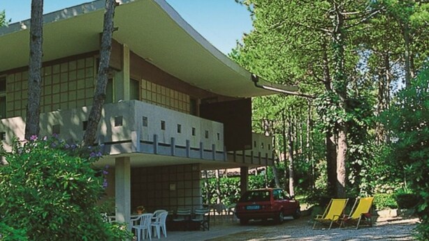 A two-story building with a flat roof and a balcony. There are trees and shrubs in front of the building. - Villa Tiziana (Lignano Sabbiadoro)