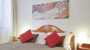 A neatly made bed with a wooden headboard, red and white pillows, and a decorative painting above it. - Grimaldi Apartments - Lionello (Venice)