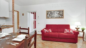 A living room with a red sofa, a dining table with chairs, and a kitchen area with a brick wall. - Grimaldi Apartments - Lionello (Venice)