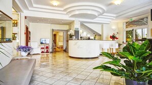 A hotel lobby with a reception desk, seating area, and a staircase leading to upper floors. - Europa (Cervia)