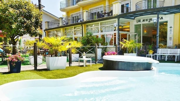 A swimming pool with a fountain, surrounded by potted plants and a building with a balcony in the background. - Europa (Cervia)