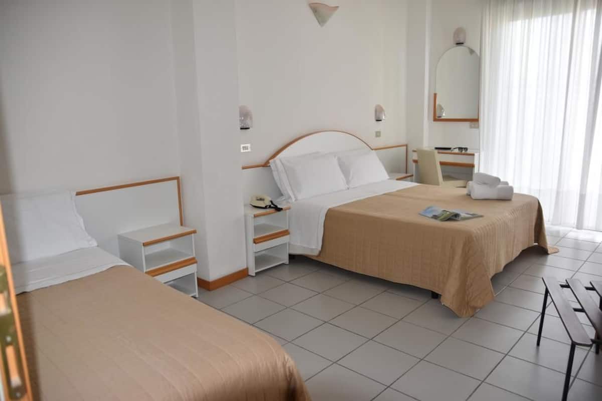 A hotel room with two beds, a bedside table with a lamp, a mirror, and a window with curtains.