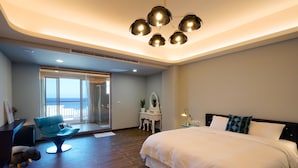 Une chambre d’hôtel moderne, dotée d’un grand lit, d’un bureau, d’une chaise et offrant une vue sur la mer grâce à une porte vitrée.