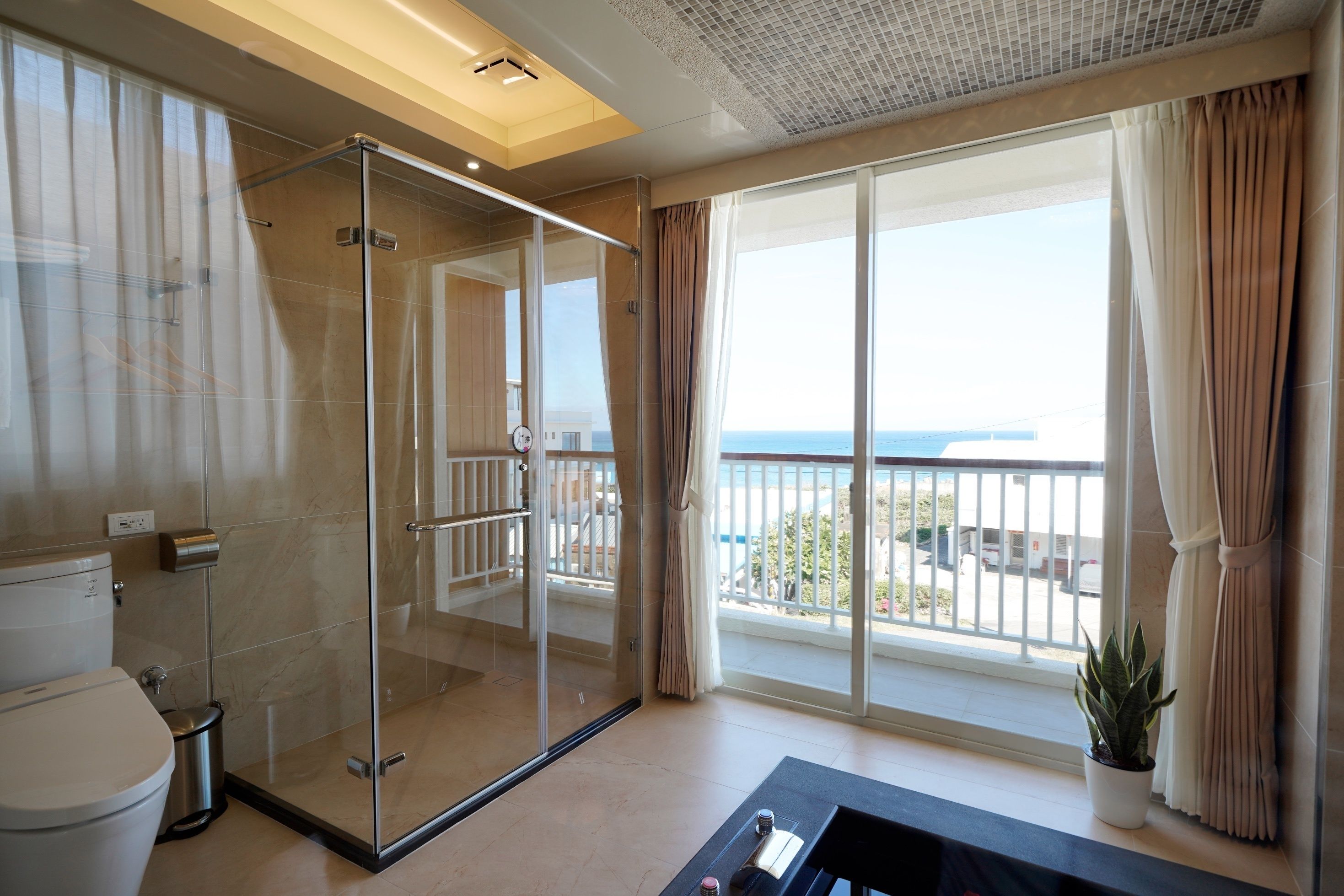 A modern bathroom with a glass shower enclosure, a toilet, and a large window with a view of the sea.