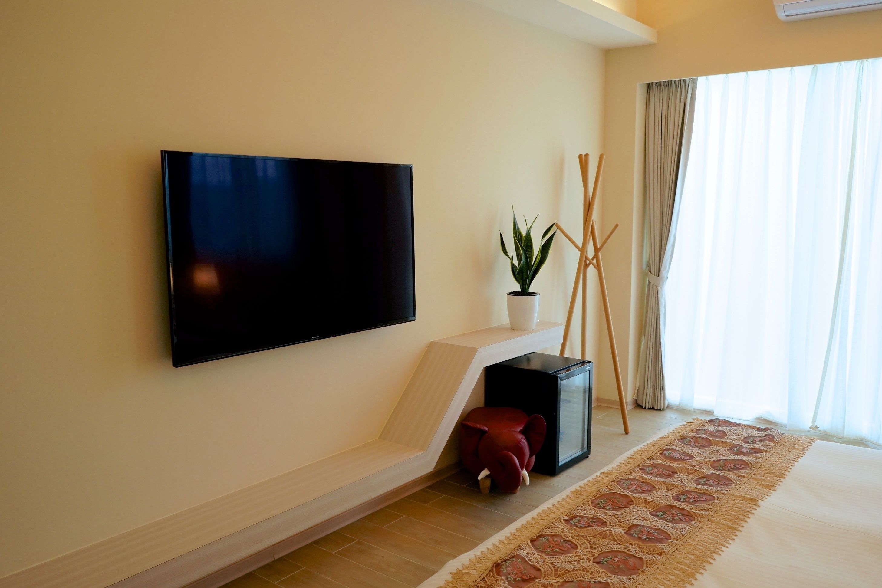 A modern hotel room with a flat-screen TV mounted on the wall, a small potted plant, a red ottoman, and a patterned rug on the floor.