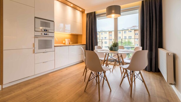 A modern kitchen with built-in appliances, a dining area with a table and chairs, and a view of buildings through large windows. - Dream Holiday Apartament (Gdansk)