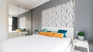A modern bedroom with a large bed, a mirror, a wall with a tree branch pattern, and a small bedside table with a plant. - Dream Holiday Apartament (Gdansk)