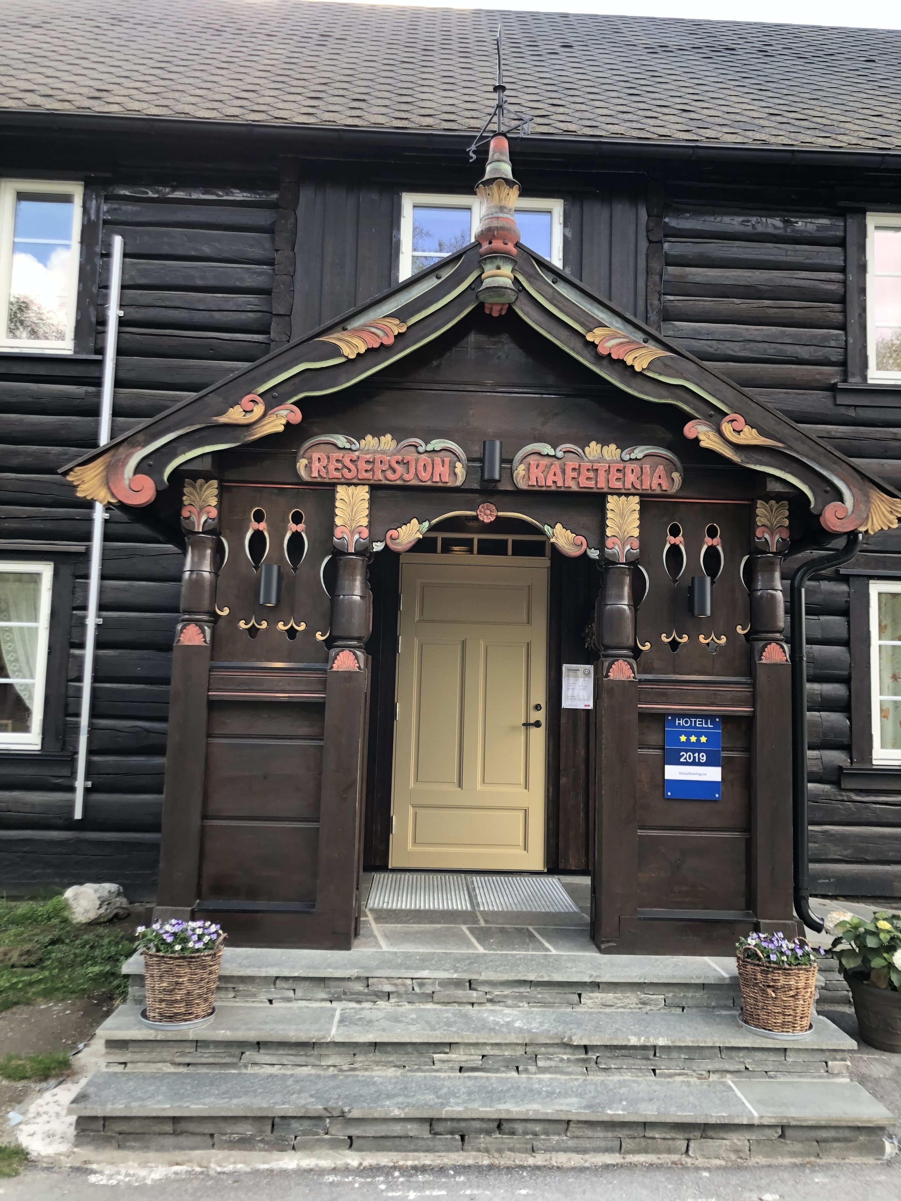 A wooden building with a sign reading "RESEPSJON KAFETERIA" and a "HOTEL" sign with a 2019 rating.