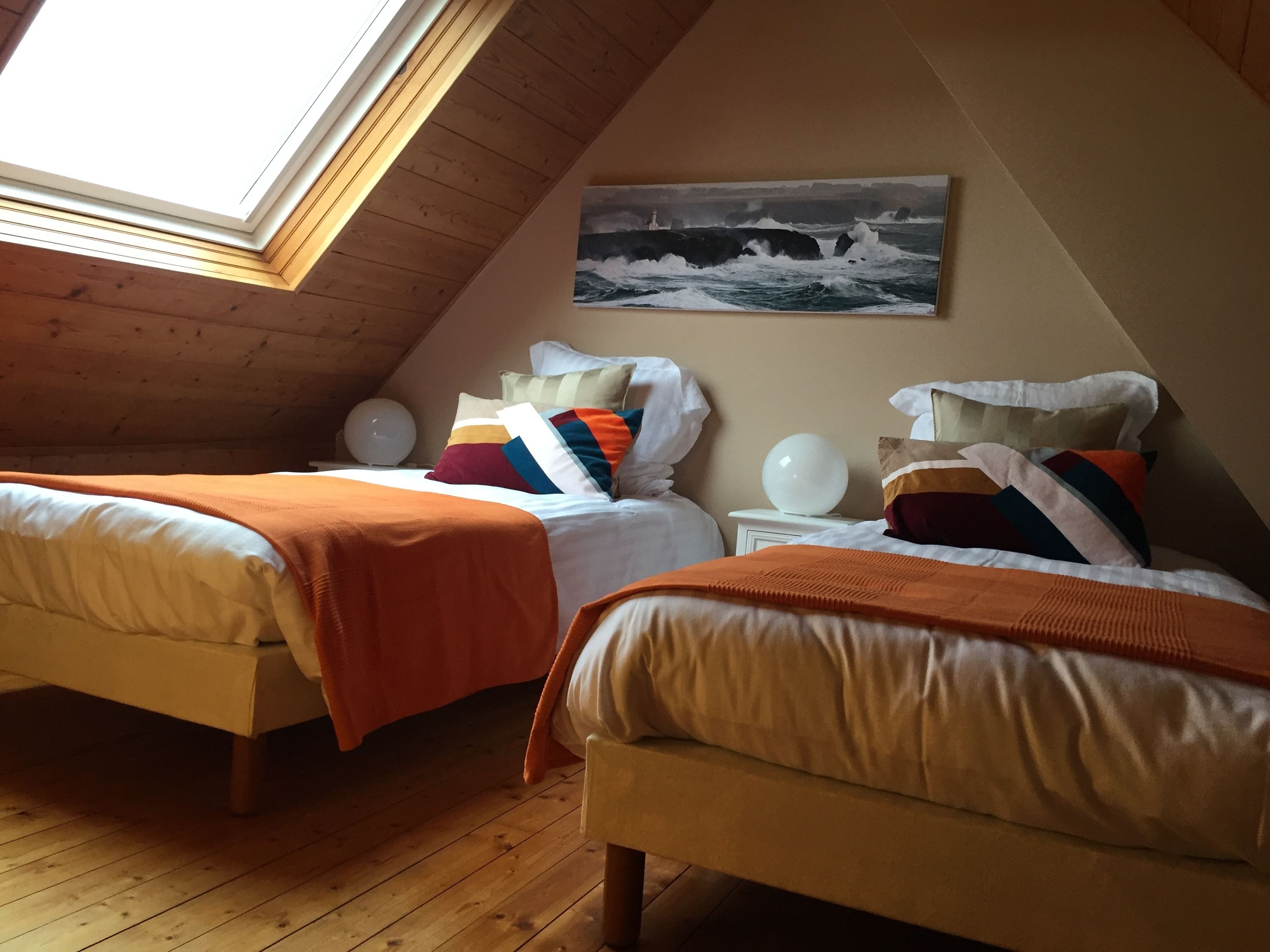 A cozy room with two beds, a painting of a lighthouse, and a skylight.