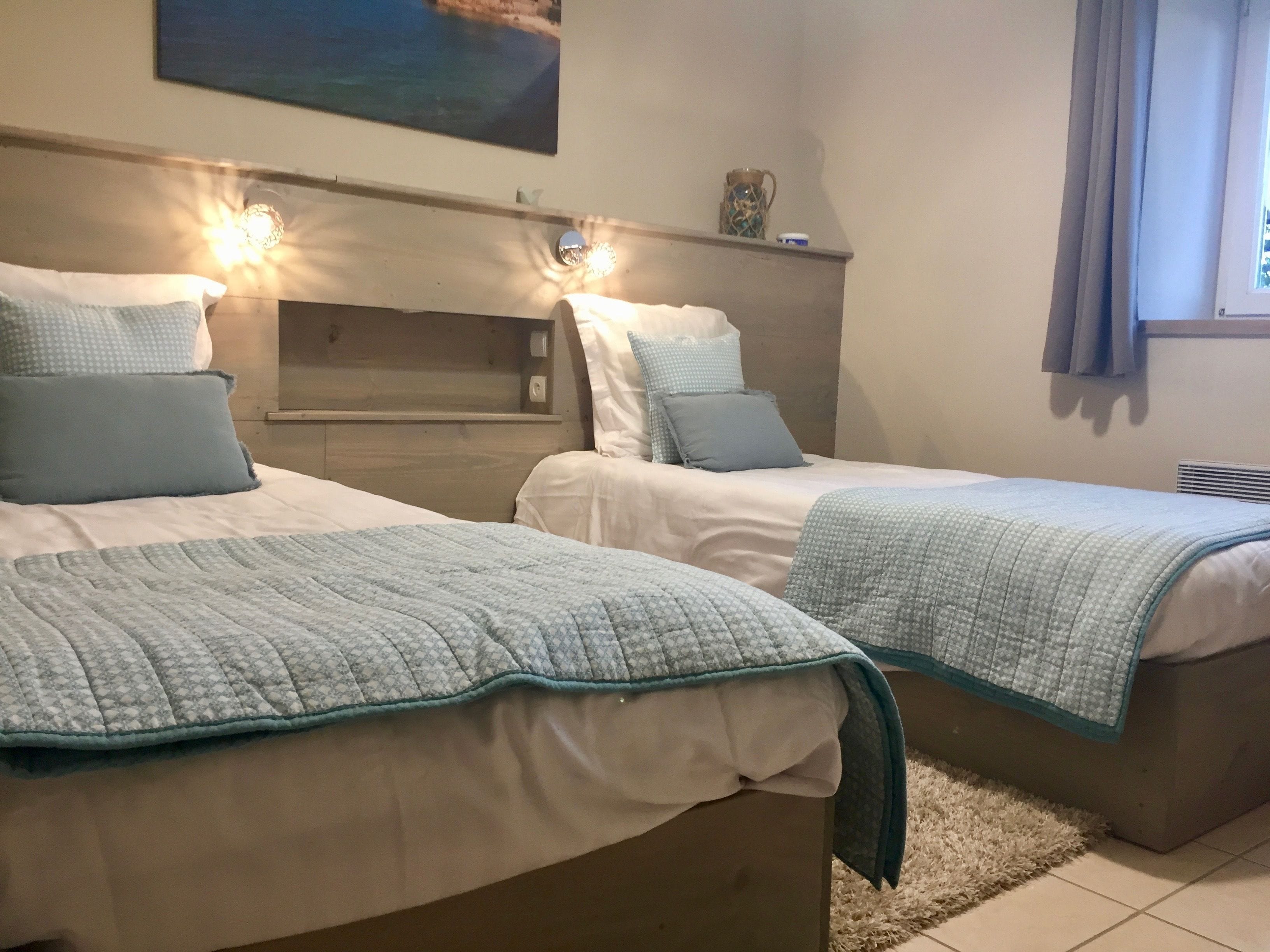 Ett hotellrum med två sängar, var och en med ett vadderat sängöverkast och kuddar. Ovanför sängarna finns väggmonterade lampor, en liten hylla med dekorativa föremål och ett fönster med gardiner.