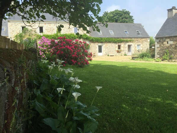 A stone building with a sloped roof, a grassy yard, and flowering plants. - Chambres d'Hotes Penker (Minihy-Treguier)