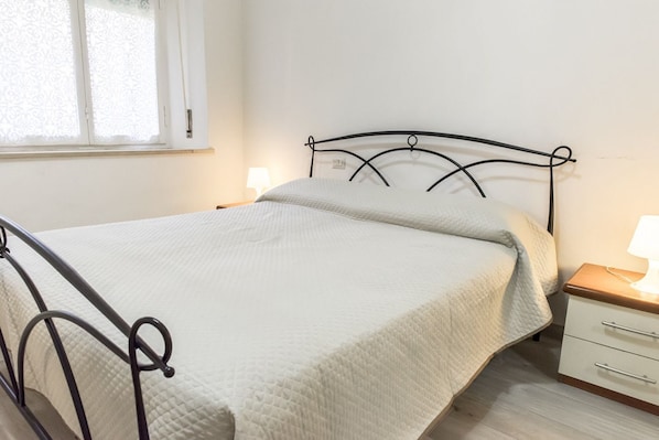 Un lit simple avec un cadre en métal, une table de chevet avec une lampe et une fenêtre avec un rideau blanc.