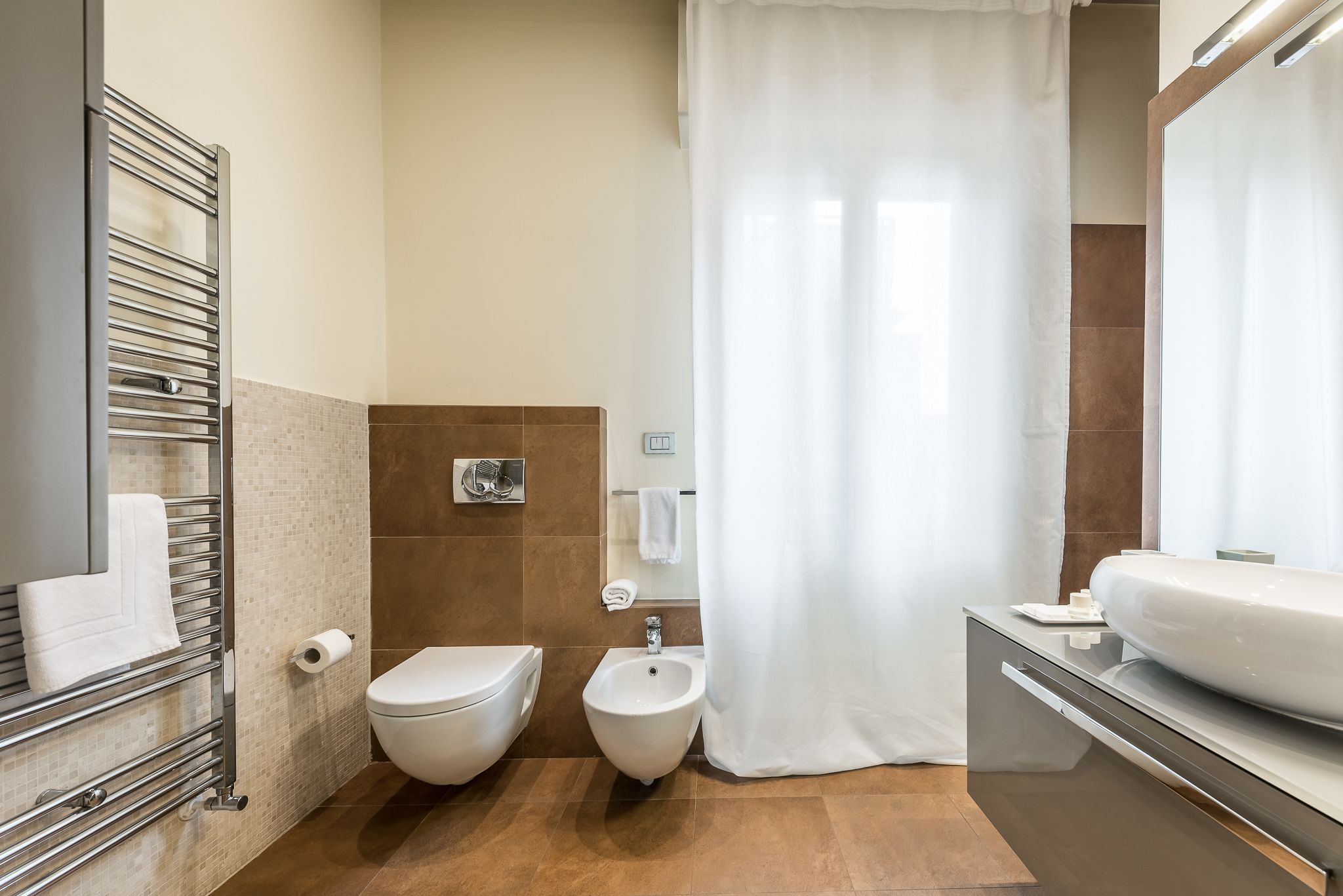 A modern bathroom with a toilet, bidet, towel rack, and a large mirror.