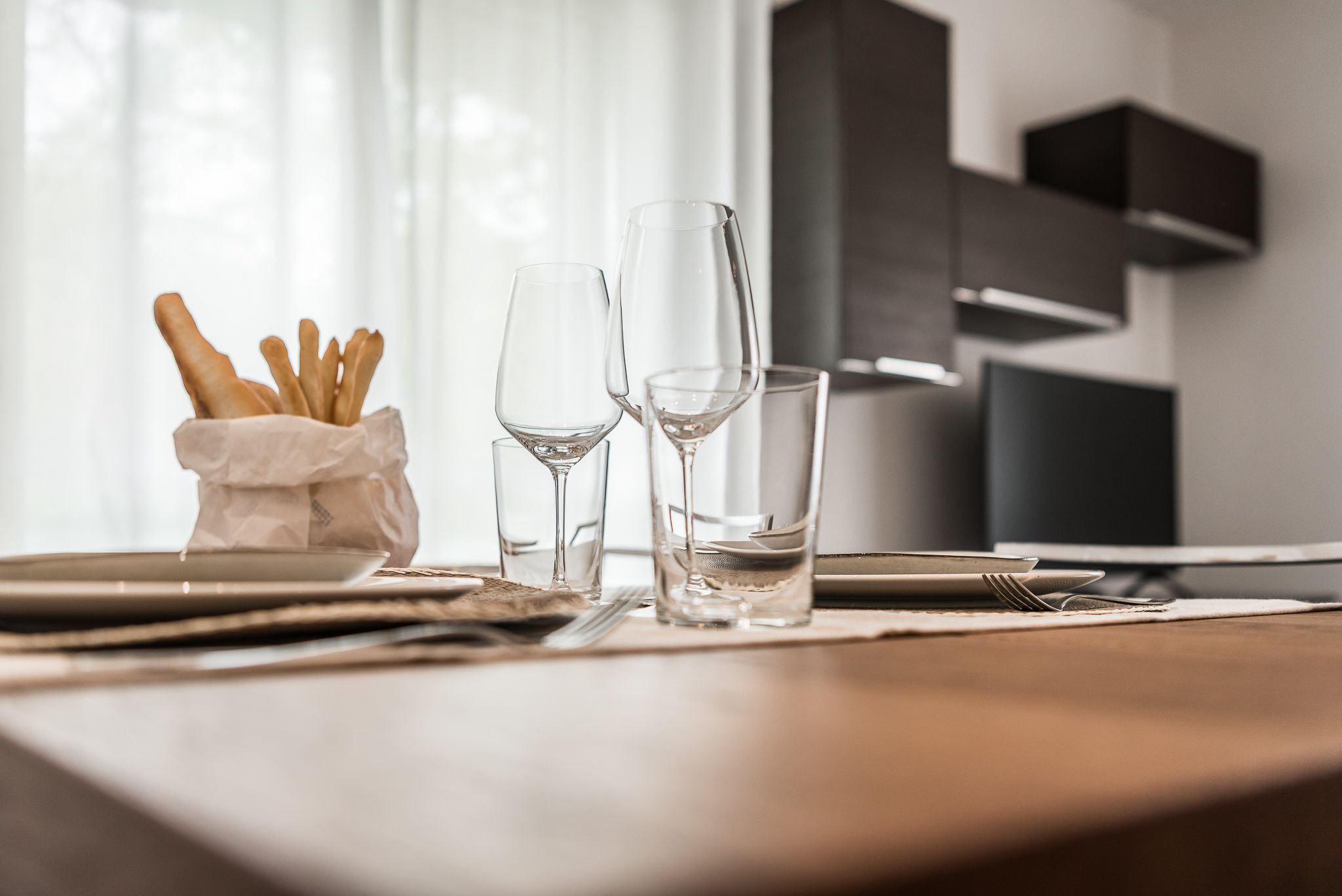 A dining table with plates, cutlery, and glasses, with a basket of breadsticks on the left.