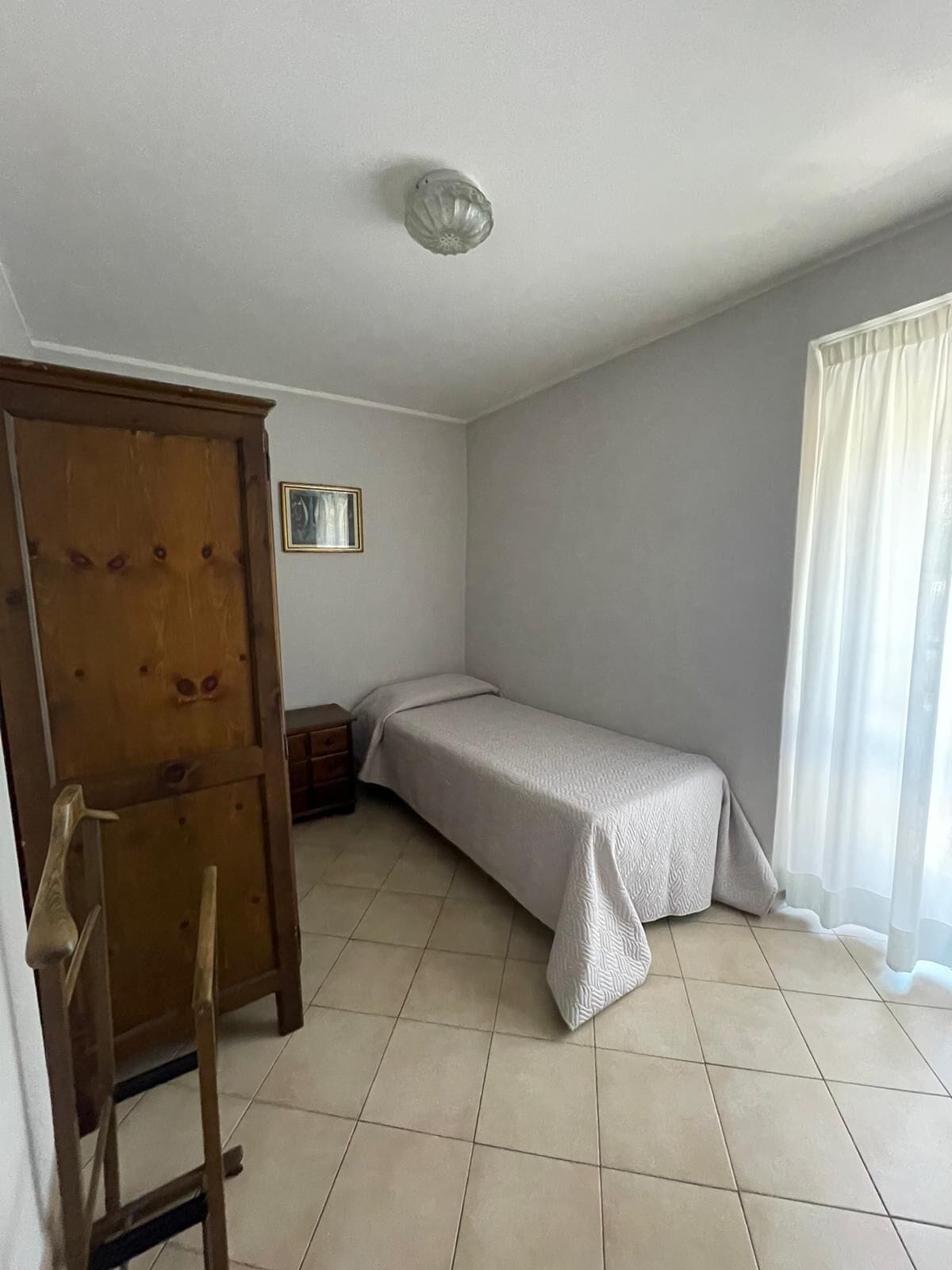 A small, sparsely furnished room with a bed, a wooden wardrobe, a small table, and a single framed picture on the wall.
