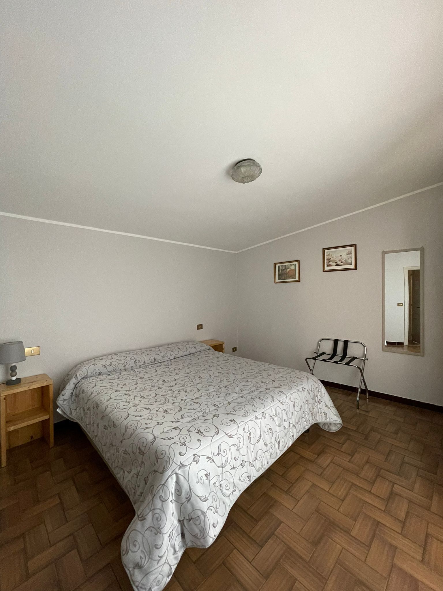 Un dormitorio con cama, mesita de noche, silla, espejo y dos cuadros en la pared.