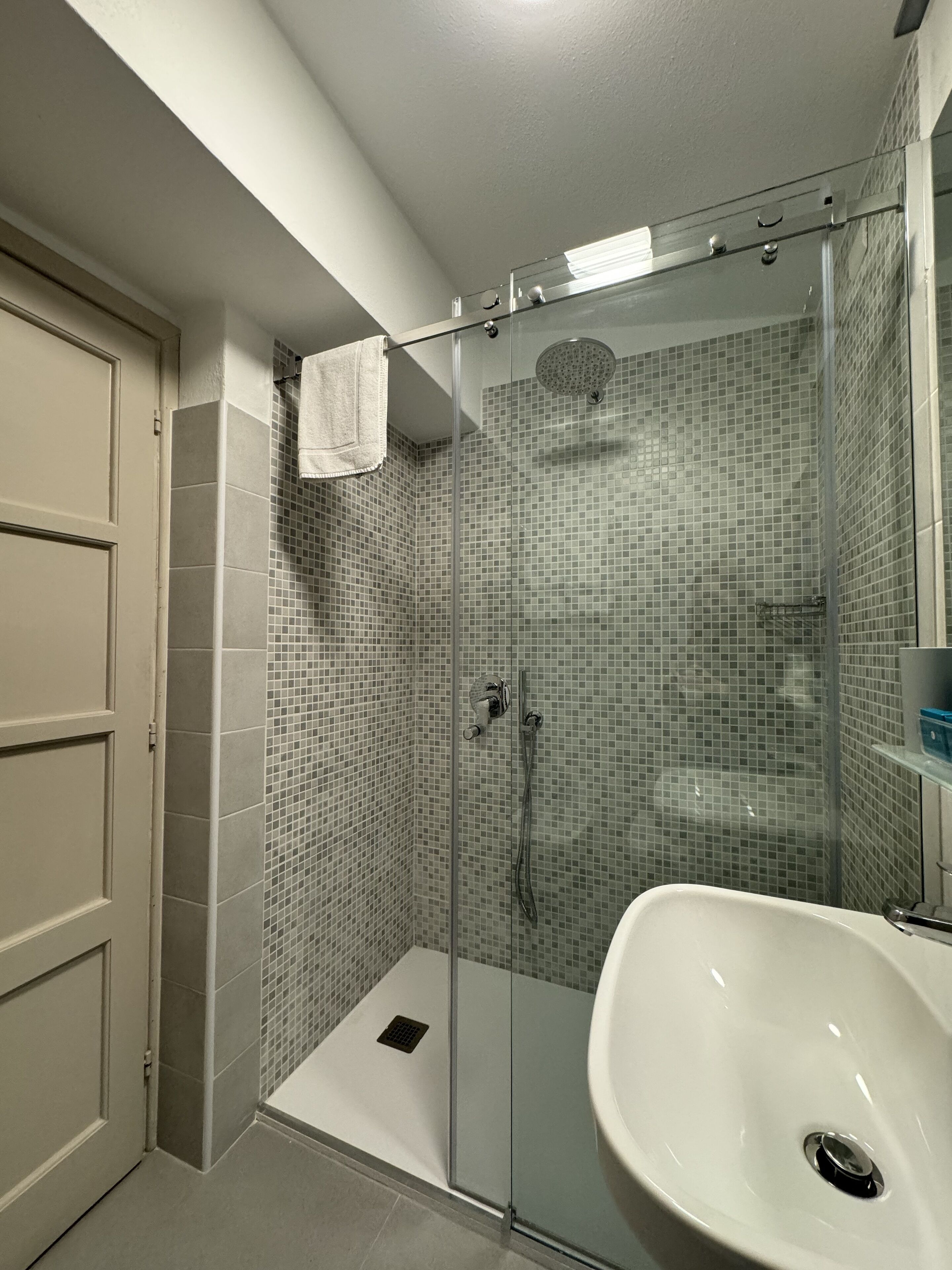 Een badkamer met een douchecabine van glas, een wit wastafeltje en een betegelde douchewand.