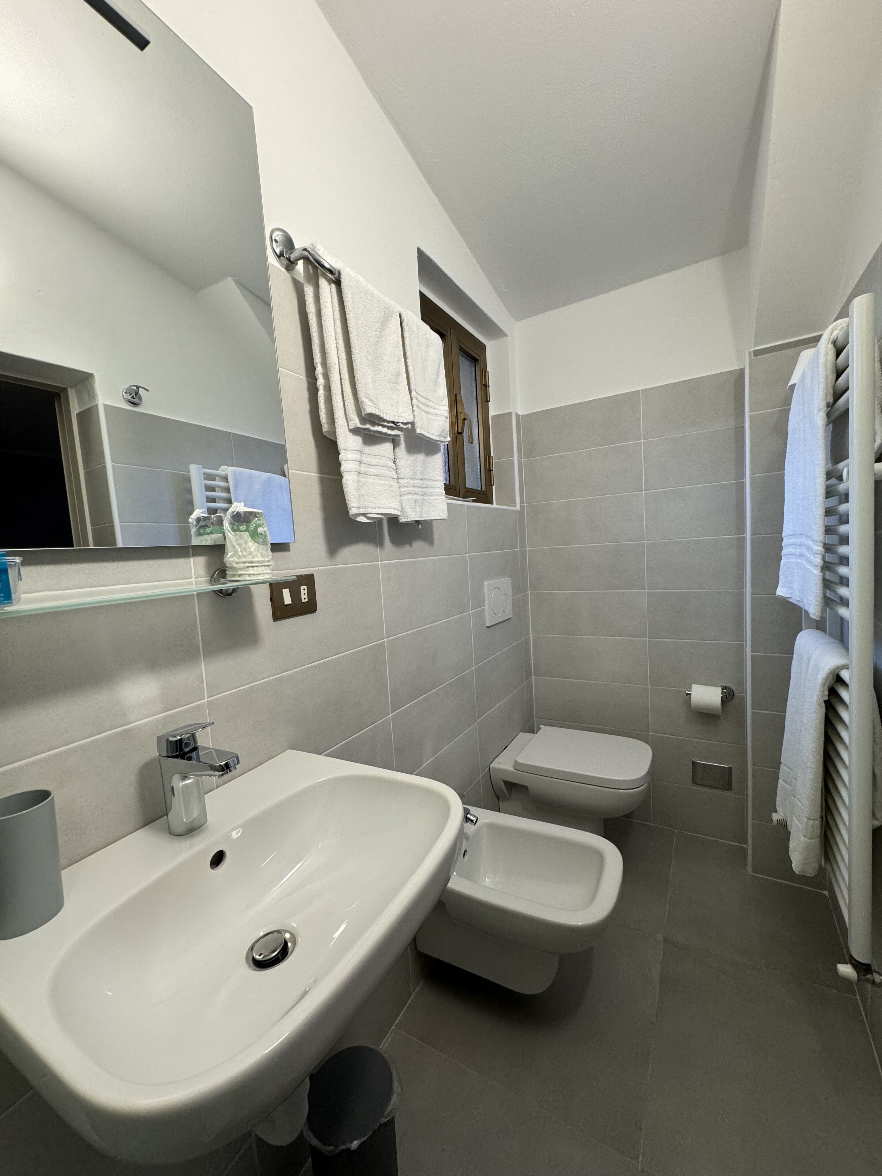 Une salle de bain moderne avec un grand lavabo, des toilettes et un bidet. Un miroir est placé au-dessus du lavabo, et des serviettes sont suspendues sur un porte-serviettes.