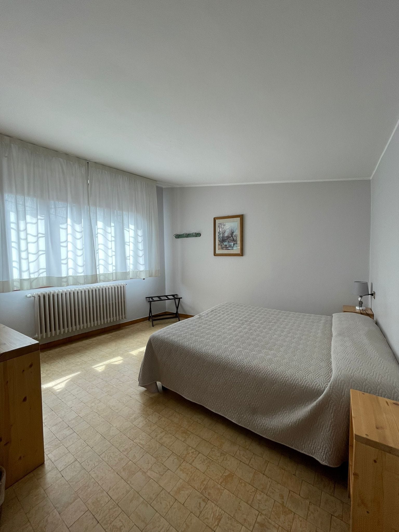 Une chambre à coucher avec un lit, un radiateur, une fenêtre avec des rideaux légers, une petite table et un tableau au mur.