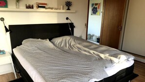 A neatly made bed with a black headboard, a bedside table with a lamp, and a shelf with books and decorative items. - Schönes Ferienhaus in Strandnähe (Rødekro)