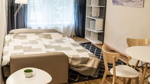 A neatly arranged hotel room with a bed, a small table with a cup, a chair, a bookshelf, and a painting on the wall. - Elizabetes Center Apartments (Riga)
