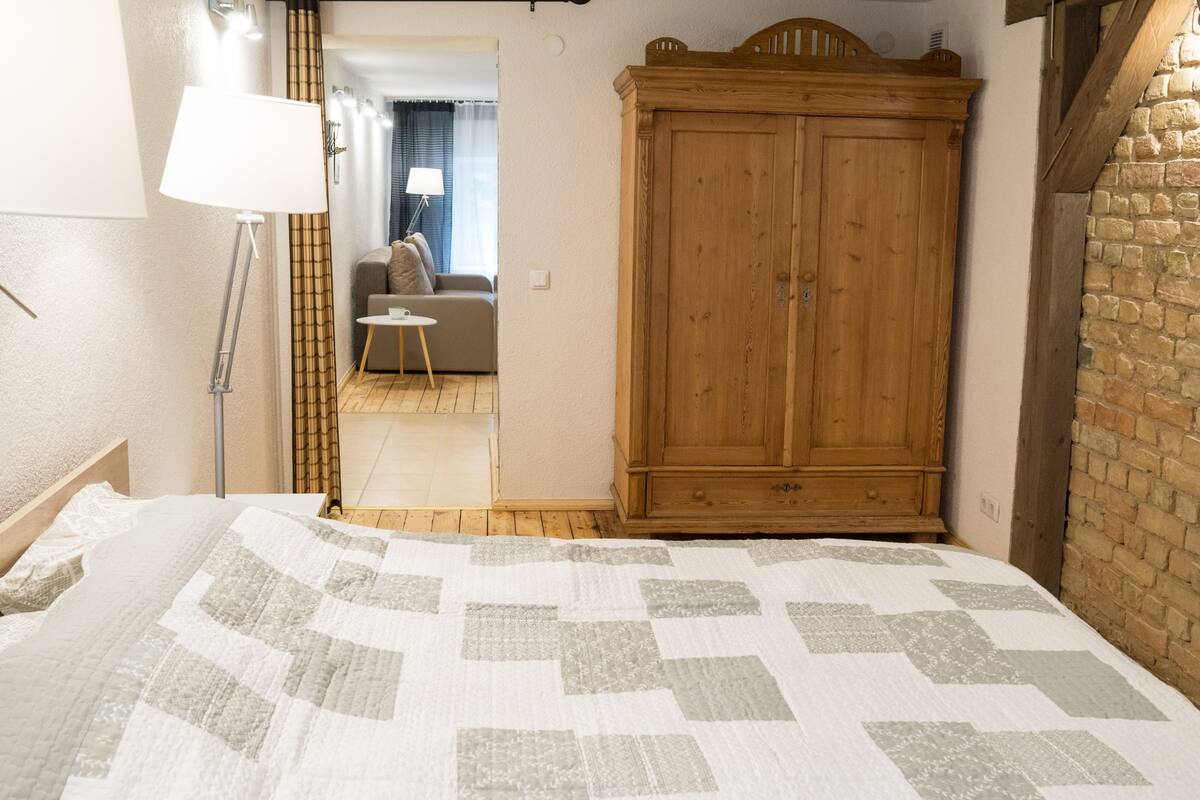A bedroom with a bed, a wooden wardrobe, a floor lamp, and a view into another room with a sofa and curtains.
