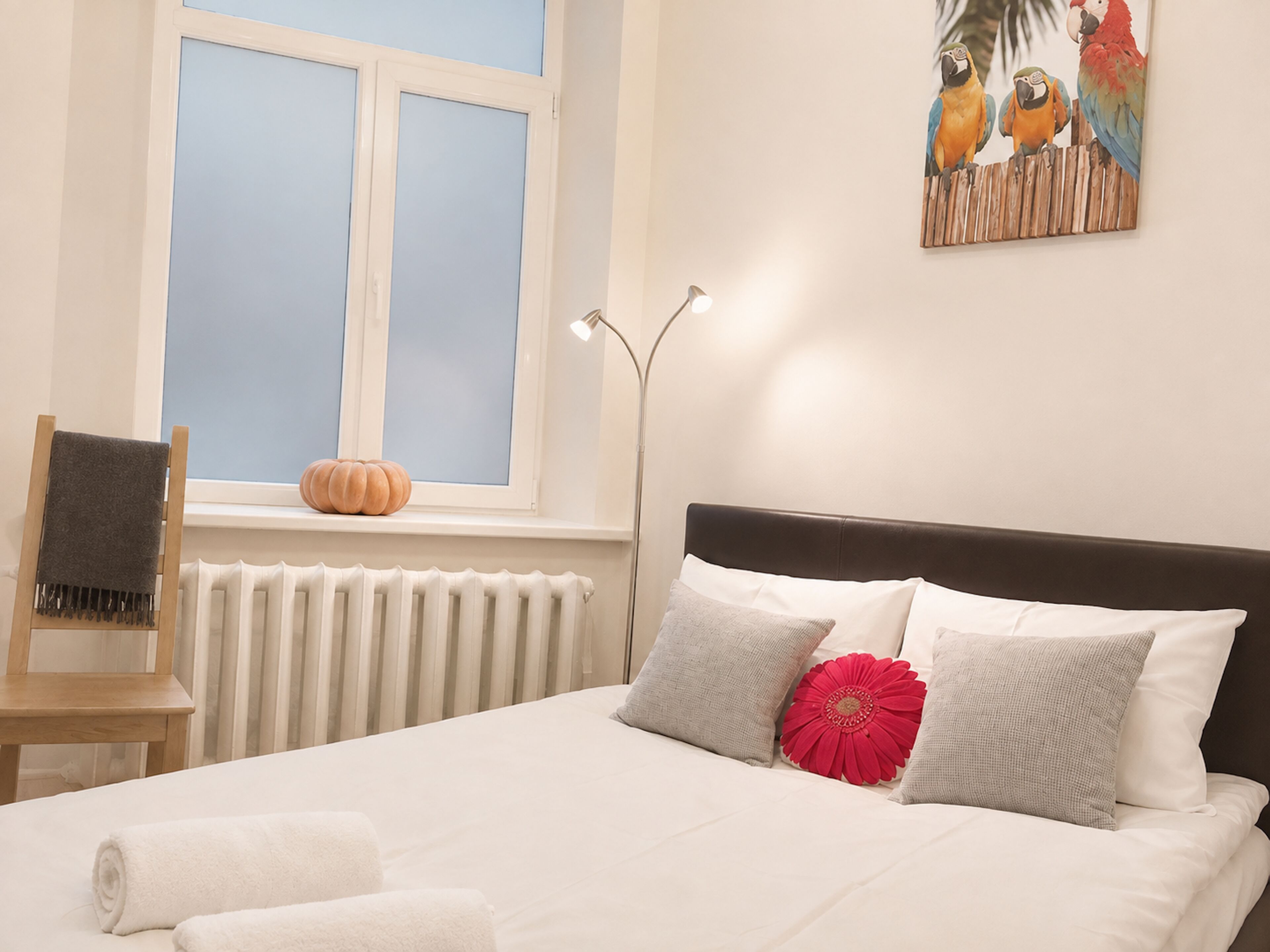 Ein Schlafzimmer mit einem Bett, einem Fenster, einem Heizkörper, einem Stuhl und einem Papageienbild.