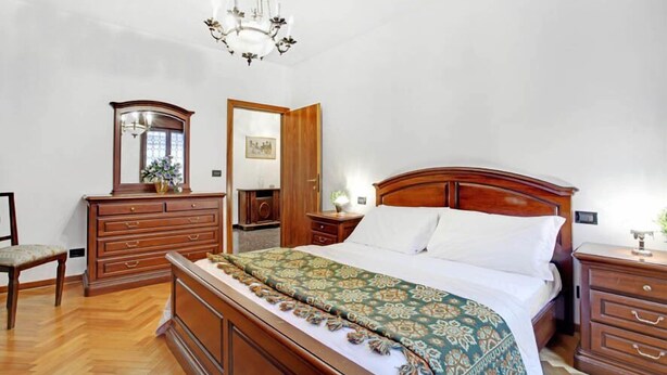 A bedroom with a wooden bed, a dresser, a chair, a mirror, and a chandelier. - Grimaldi Apartments - Elena (Venice)