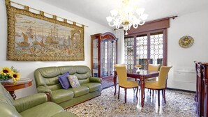 A living room with a green sofa, a dining table with chairs, a chandelier, and a large painting on the wall. - Grimaldi Apartments - Elena (Venice)