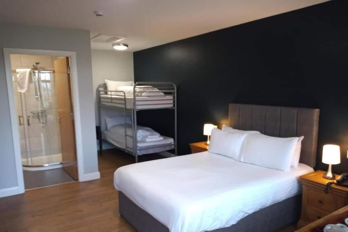A hotel room with a large bed, a bedside table with a lamp, a bunk bed, and a glass shower enclosure.