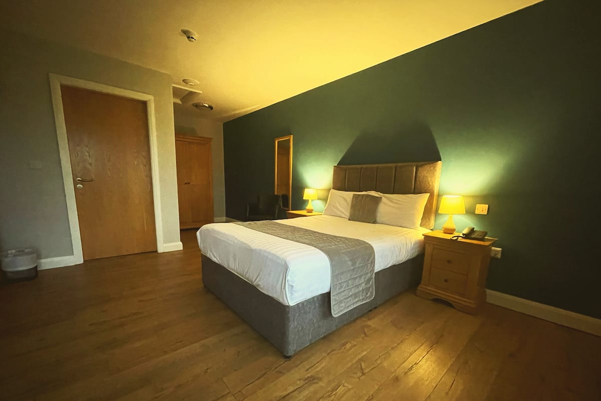 A bedroom with a large bed, wooden floor, two bedside tables with lamps, and a door leading to another room.