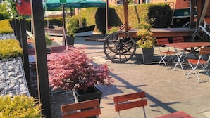 An outdoor seating area with wooden benches and tables, a decorative wheel, and potted plants. - Rüstersieler Hof (Wilhelmshaven)