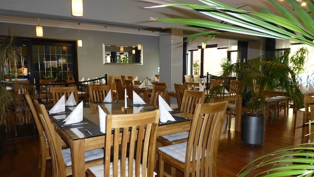 A restaurant with wooden tables and chairs, a large potted plant, and a modern interior design. - Rüstersieler Hof (Wilhelmshaven)