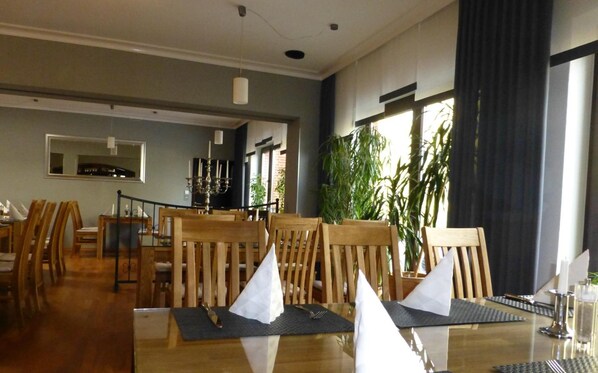 A dining area with wooden chairs and tables, a chandelier, and large windows with curtains. - Rüstersieler Hof (Wilhelmshaven)