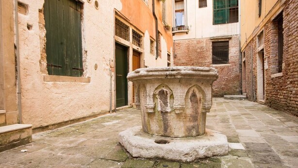 A narrow alleyway with a stone fountain in the center, flanked by buildings with arched windows and doors. - Grimaldi Apartments - San Marco Economy (Venice)