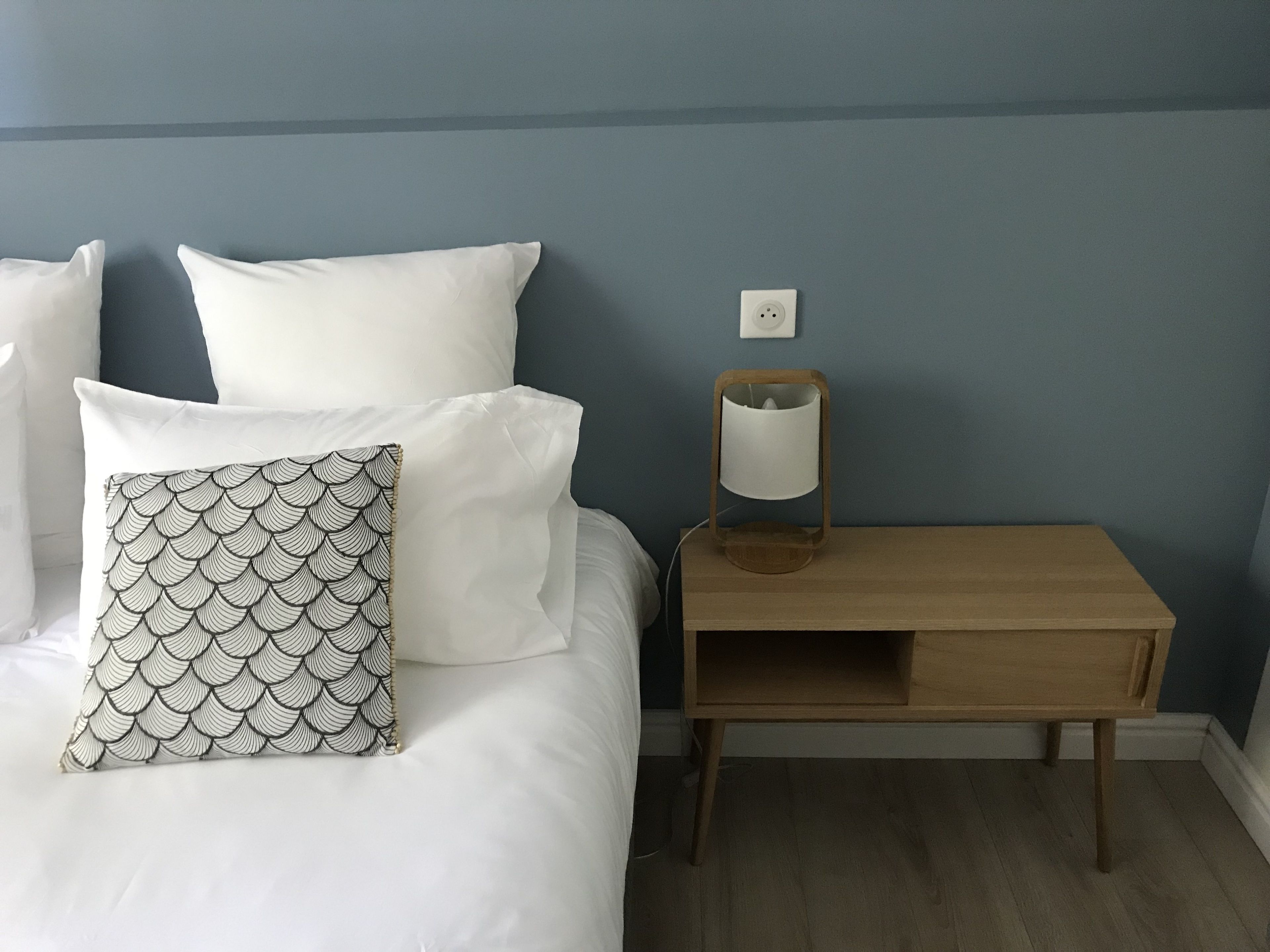 Un lit bien fait, avec du linge de lit blanc et un coussin à motifs, une table de chevet avec une lampe et une prise électrique sur le mur.