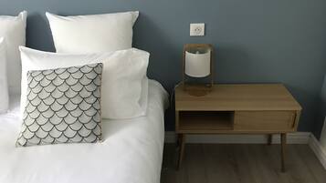 Un lit bien fait, avec du linge de lit blanc et un coussin à motifs, une table de chevet avec une lampe et une prise électrique sur le mur.