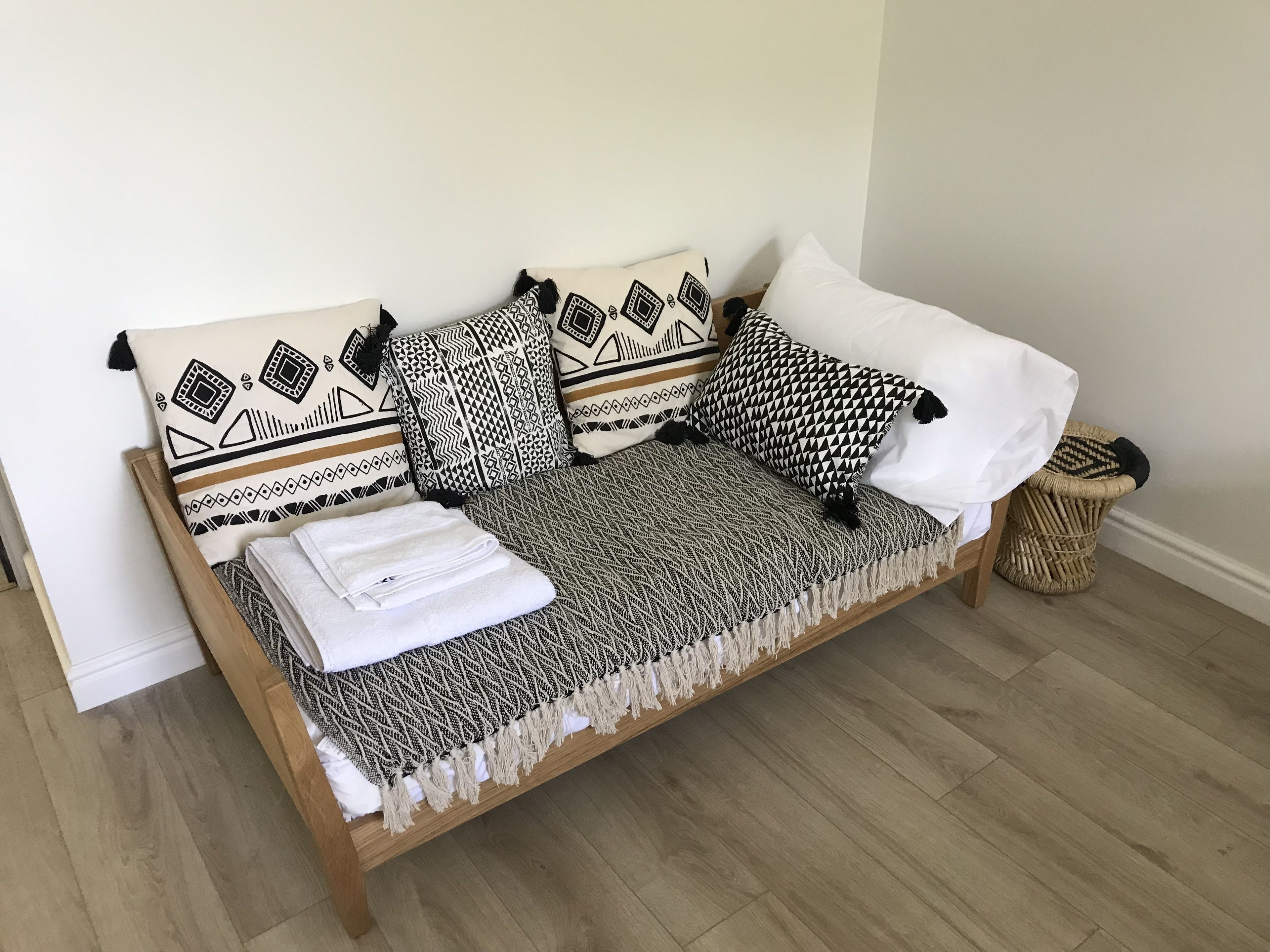 A wooden sofa bed with patterned cushions, a folded white towel, and a woven basket.