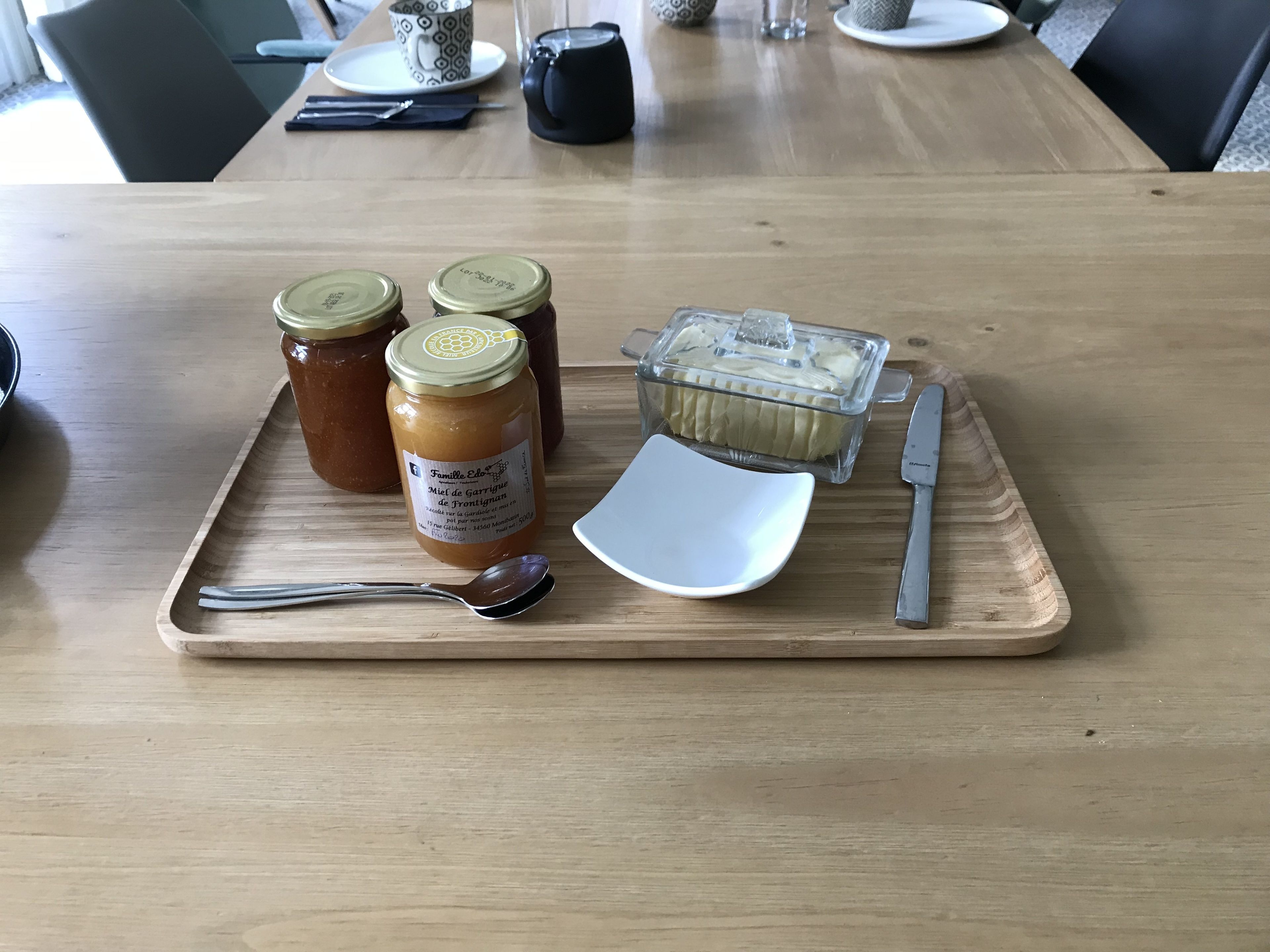 A wooden table with a tray holding two jars of jam, a container of cream, a spoon, and a knife.