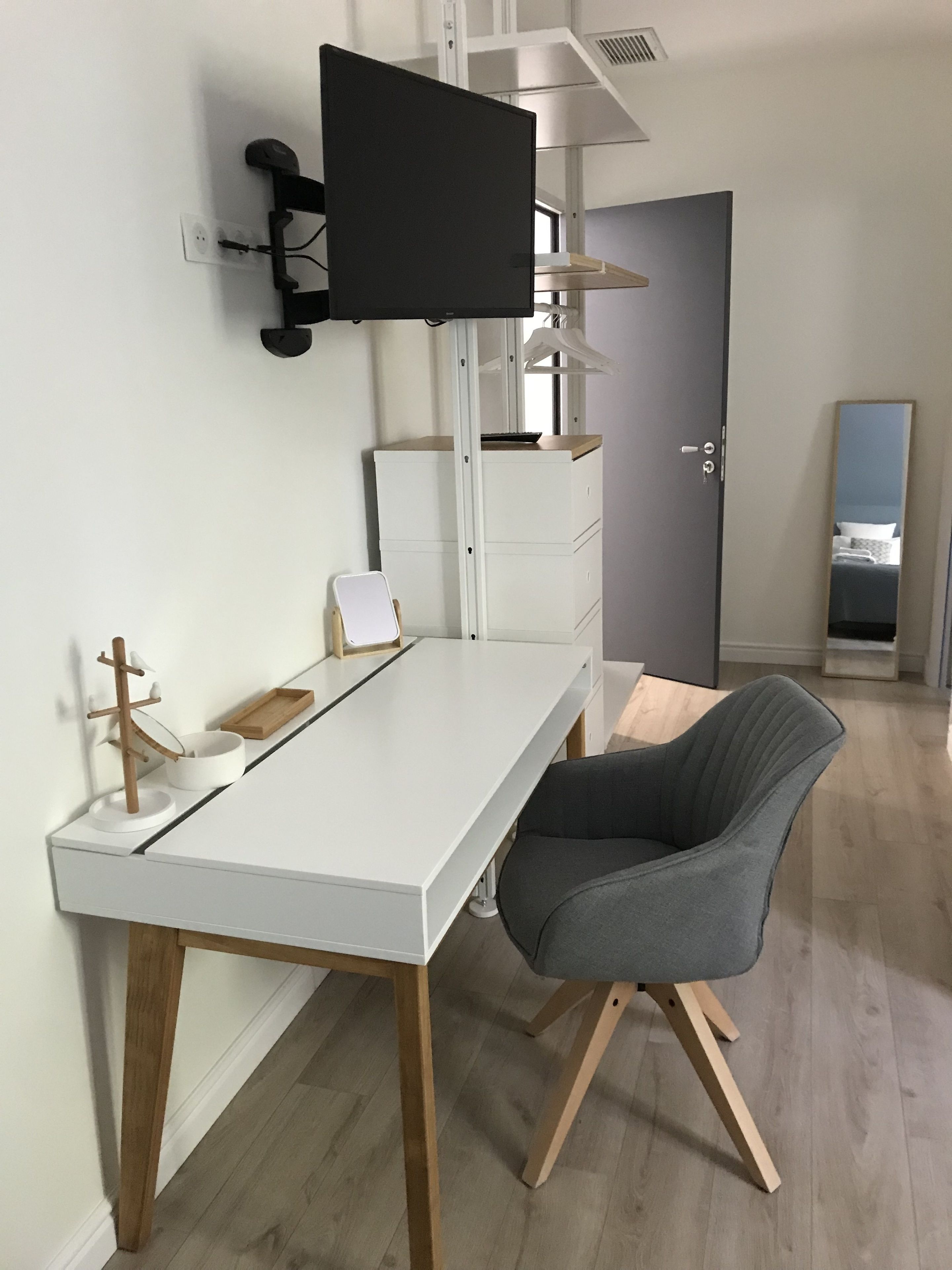A modern room with a white desk, a grey chair, a flat-screen TV mounted on the wall, and a wooden floor.