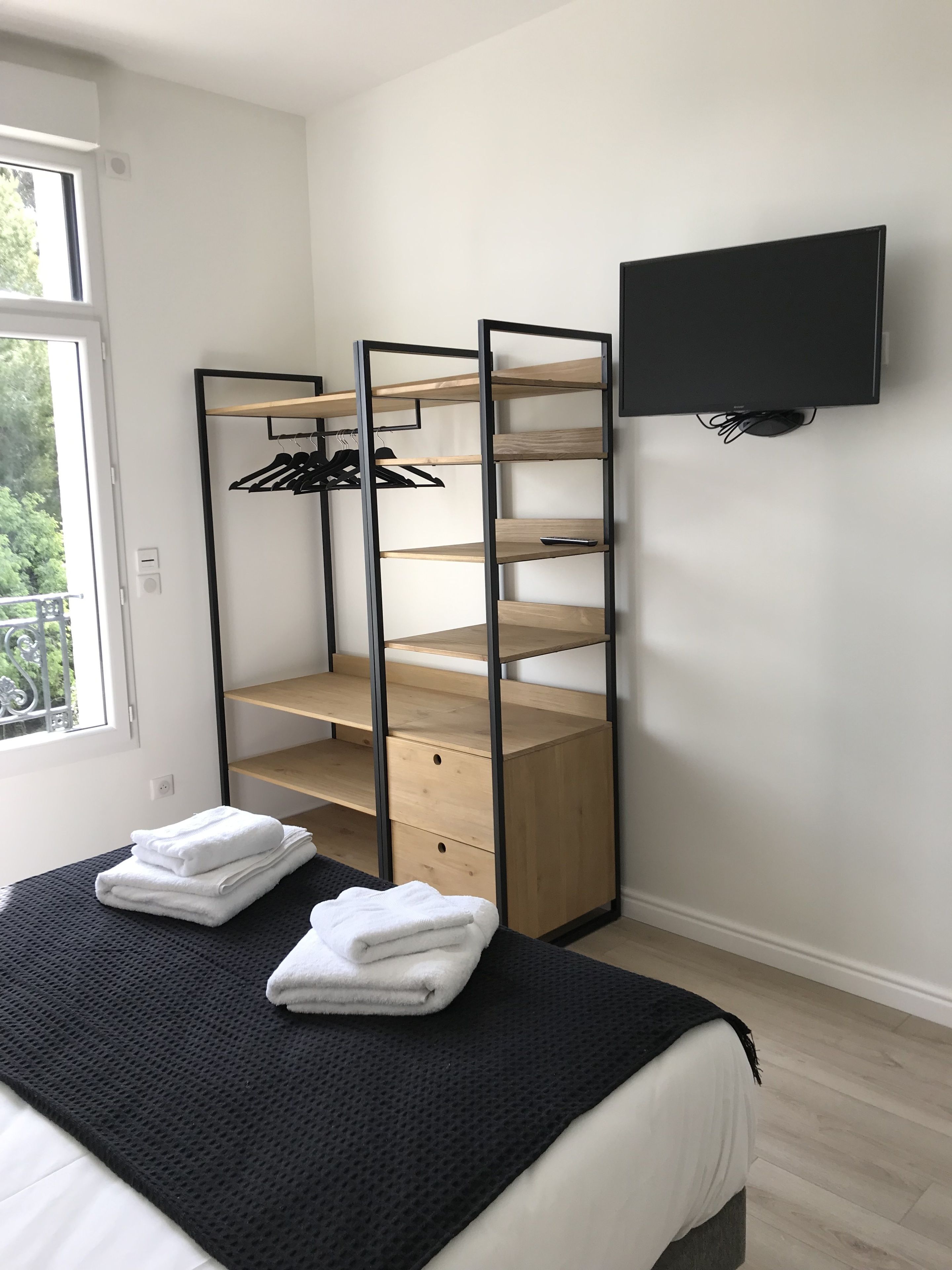 Quarto com cama, televisão preta fixada na parede e estante de madeira com roupas.