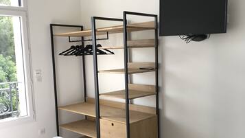 Quarto com cama, televisão preta fixada na parede e estante de madeira com roupas.