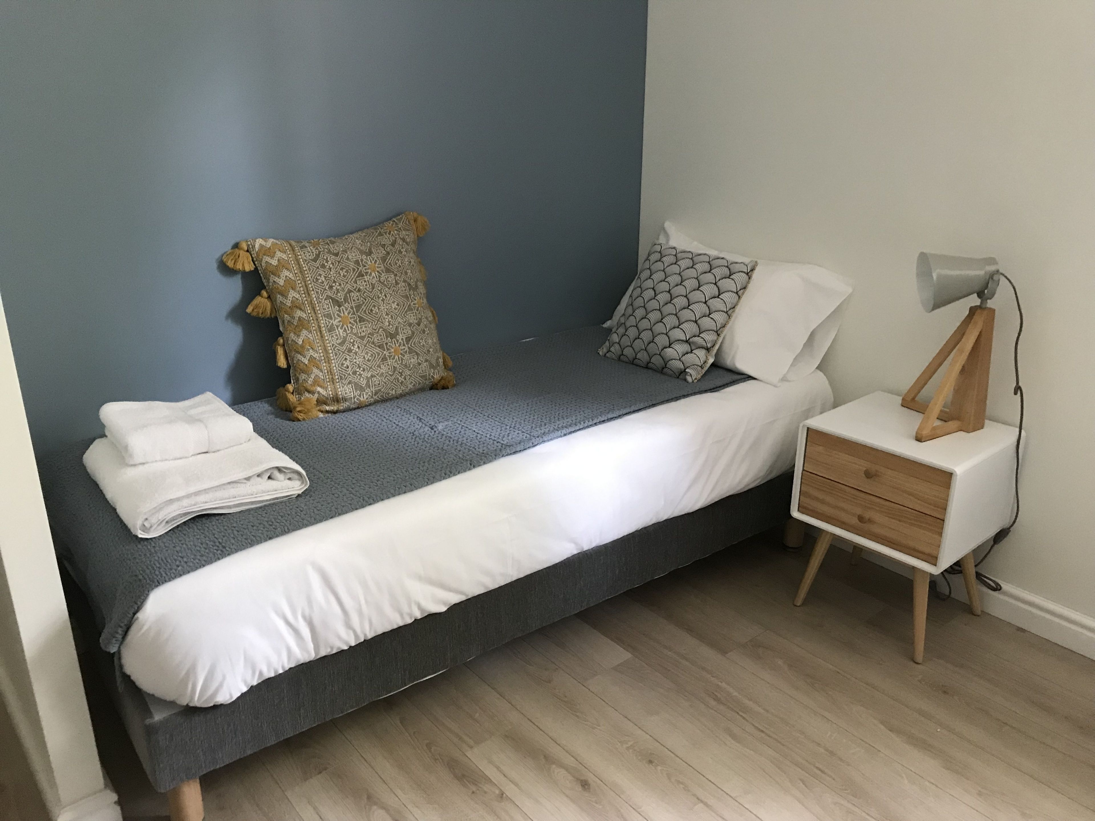 Una cama individual con colchón gris, almohadas blancas y una almohada decorativa estampada. Se observa una mesita de noche con una lámpara y una toalla blanca doblada.