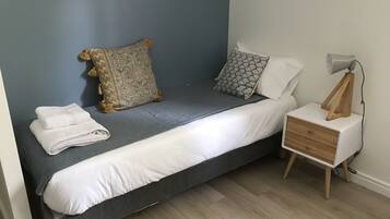 Una cama individual con colchón gris, almohadas blancas y una almohada decorativa estampada. Se observa una mesita de noche con una lámpara y una toalla blanca doblada.