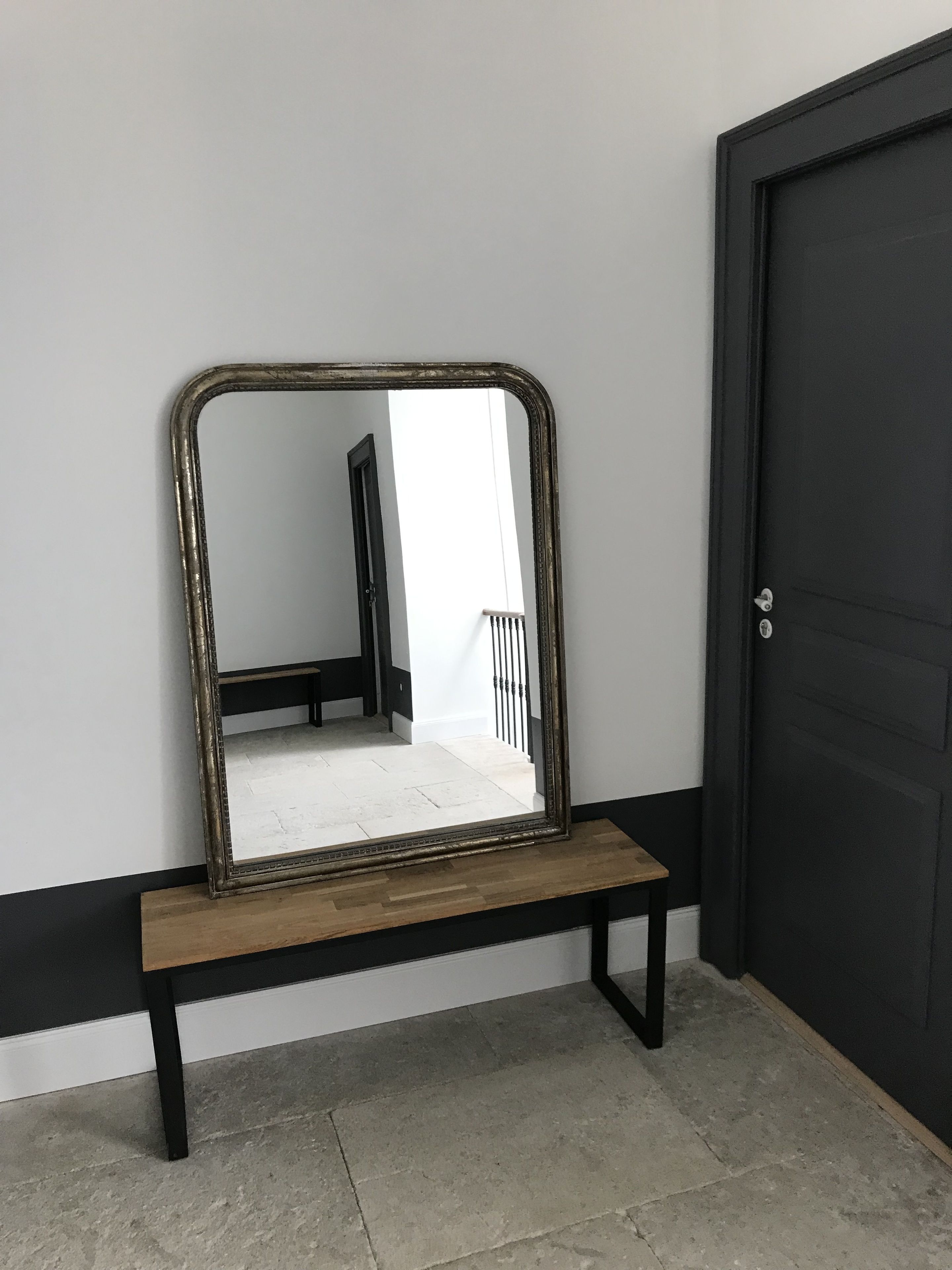 Un miroir posé sur un support en bois, adossé à un mur, avec une porte sur la droite.