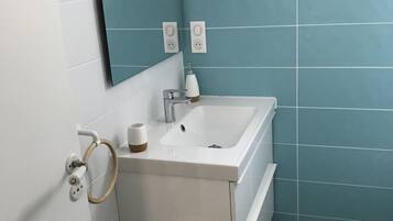 Un baño con un lavamanos blanco, un espejo y un inodoro. Hay dos enchufes en la pared.