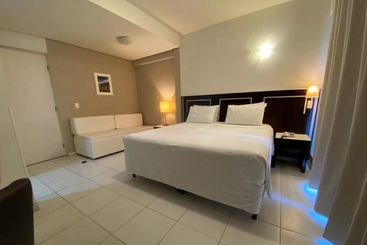 A hotel room with a large bed, a bedside table, a nightstand, a sofa, and a wall-mounted picture.