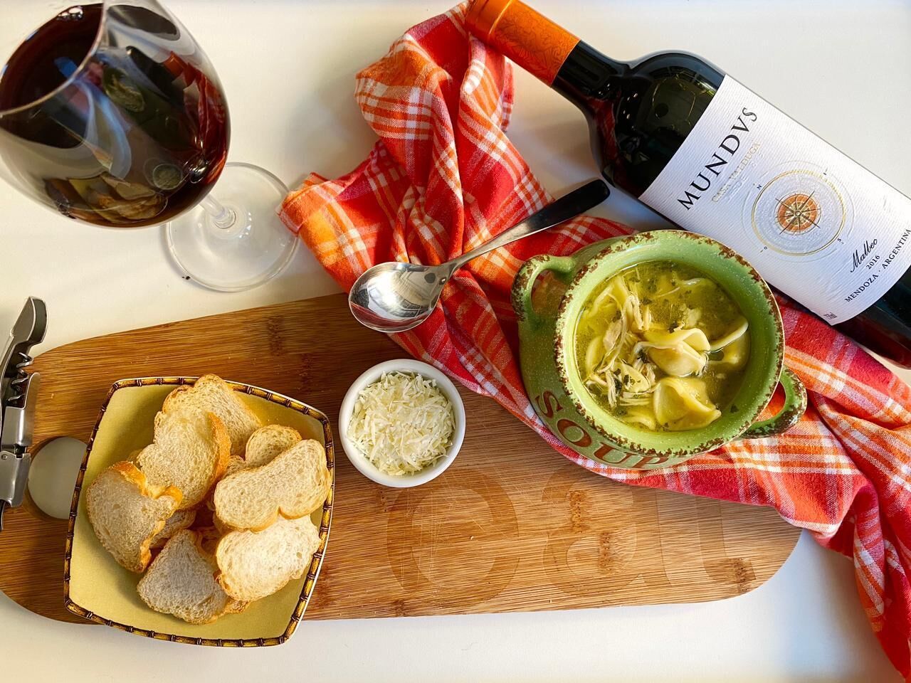 A wooden board with bread, a bowl of soup, a wine bottle, and a wine glass.