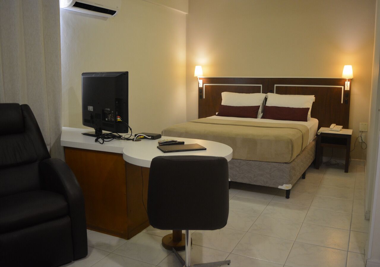 A hotel room with a bed, a desk with a computer, a chair, and a bedside table with a lamp.