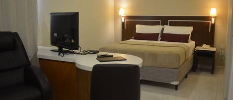A hotel room with a bed, a desk with a computer, a chair, and a bedside table with a lamp.